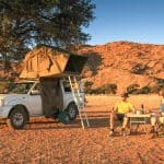 Motor Hills | How To Convert a Truck Bed Into a Camper (Ultimate Guide) Happy African American couple having dinner at sunset next to a pickup truck with a tent on top of it with a ladder