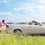 A man pushing a broken down convertible car on the side of the road to push start it while the girl is behind the wheel
