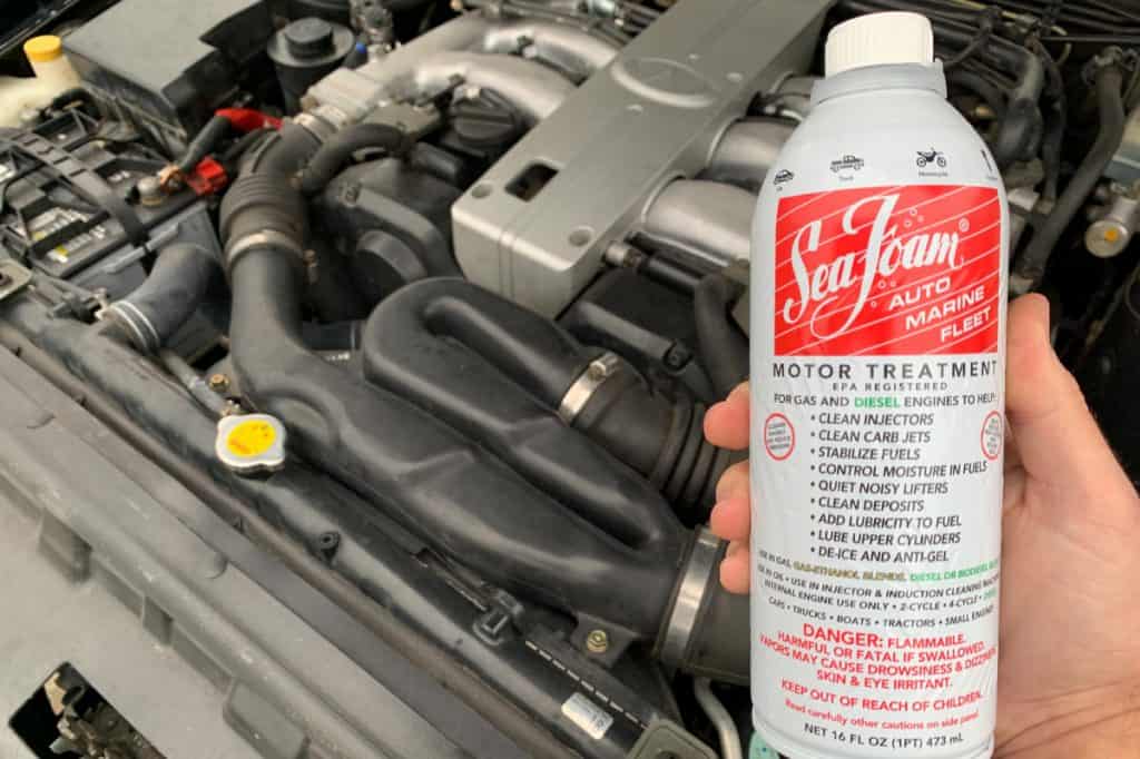 Motor Hills | Is Seafoam Good For Your Engine? Seafoam bottle being held up against a car engine of an Infiniti J30 in the background